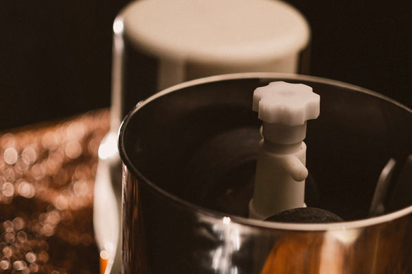 A close-up shot of the Spectra 11 Chocolate Melanger, featuring a white base, stainless steel bowl, and orange power button, set against a bed of cacao beans with warm, moody lighting.