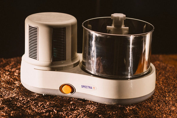 A close-up shot of the Spectra 11 Chocolate Melanger, featuring a white base, stainless steel bowl, and orange power button, set against a bed of cacao beans with warm, moody lighting.
