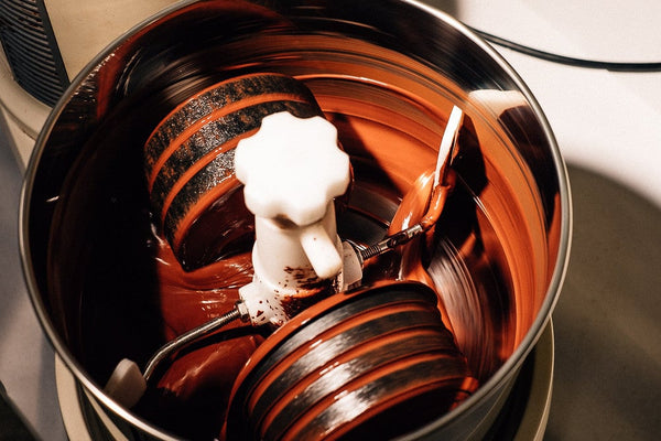 A close-up shot of glossy, dark chocolate swirling in a stainless steel melanger drum, with a white paddle attachment partially coated in chocolate. The warm lighting highlights the texture and sheen of the melted chocolate.