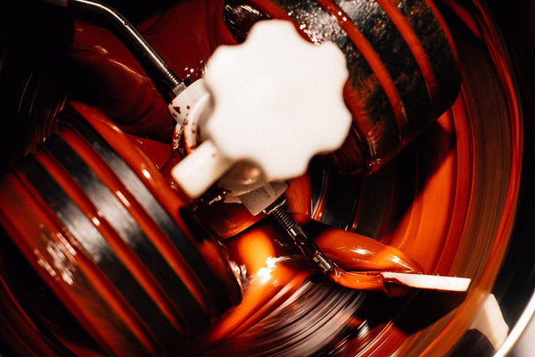 A close-up shot of glossy, dark chocolate swirling in a stainless steel melanger drum, with a white paddle attachment partially coated in chocolate. The warm lighting highlights the texture and sheen of the melted chocolate.