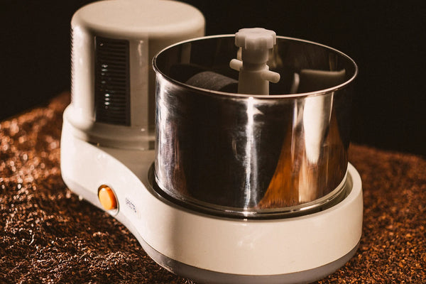 A close-up shot of the Spectra 11 Chocolate Melanger, featuring a white base, stainless steel bowl, and orange power button, set against a bed of cacao beans with warm, moody lighting.