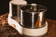A close-up shot of the Spectra 11 Chocolate Melanger, featuring a white base, stainless steel bowl, and orange power button, set against a bed of cacao beans with warm, moody lighting.